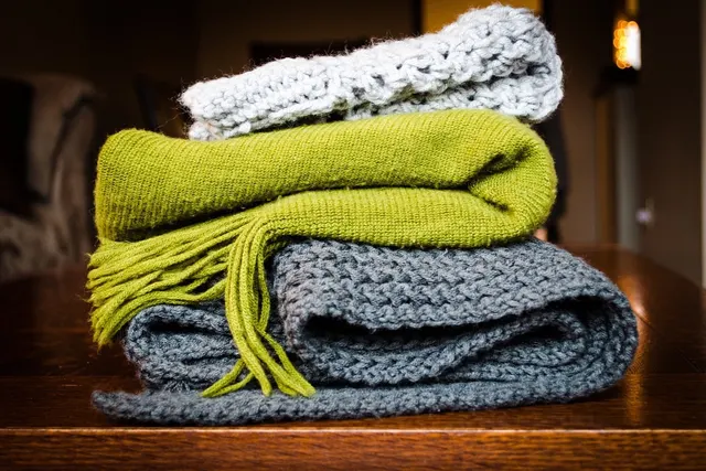 Three folded knitted throws stacked on a wooden table, a lime green fringed knit between a grey chunky blanket and a pale open-weave throw.