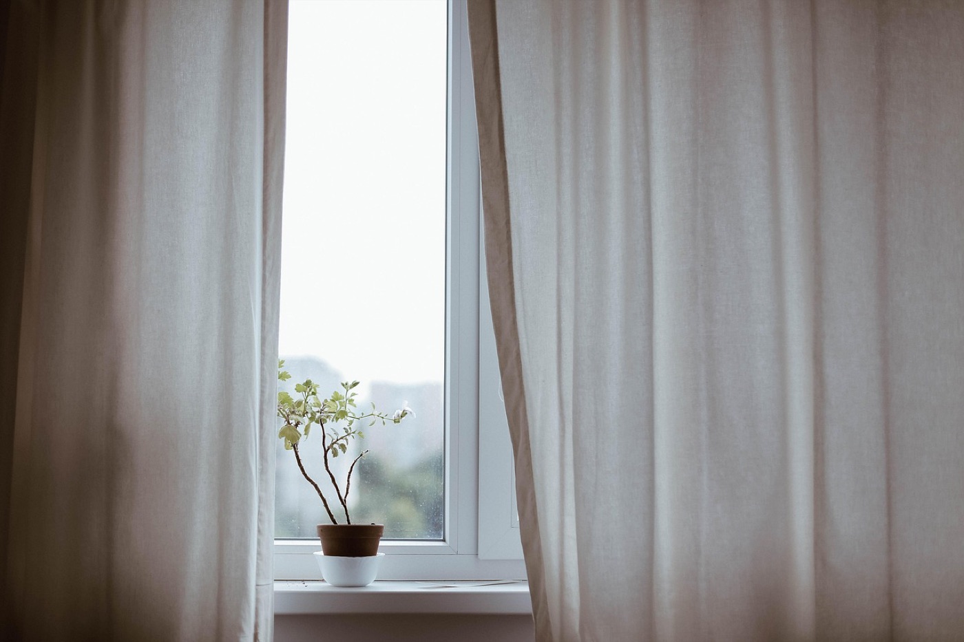 Linen curtains hung from a rod beside a tall window, with the fabric folded back and a small potted plant on the sill.