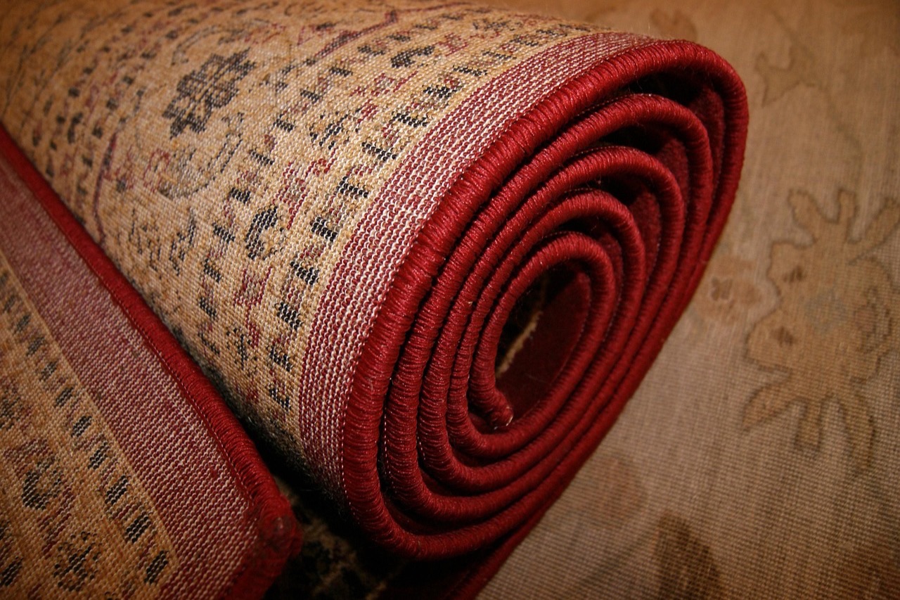 A patterned rug with a red bound edge rolled tightly, viewed from the end of the roll against a plain floor.